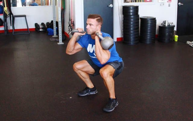 Un homme réalise un front squat avec deux kettlebells dans une salle de sport