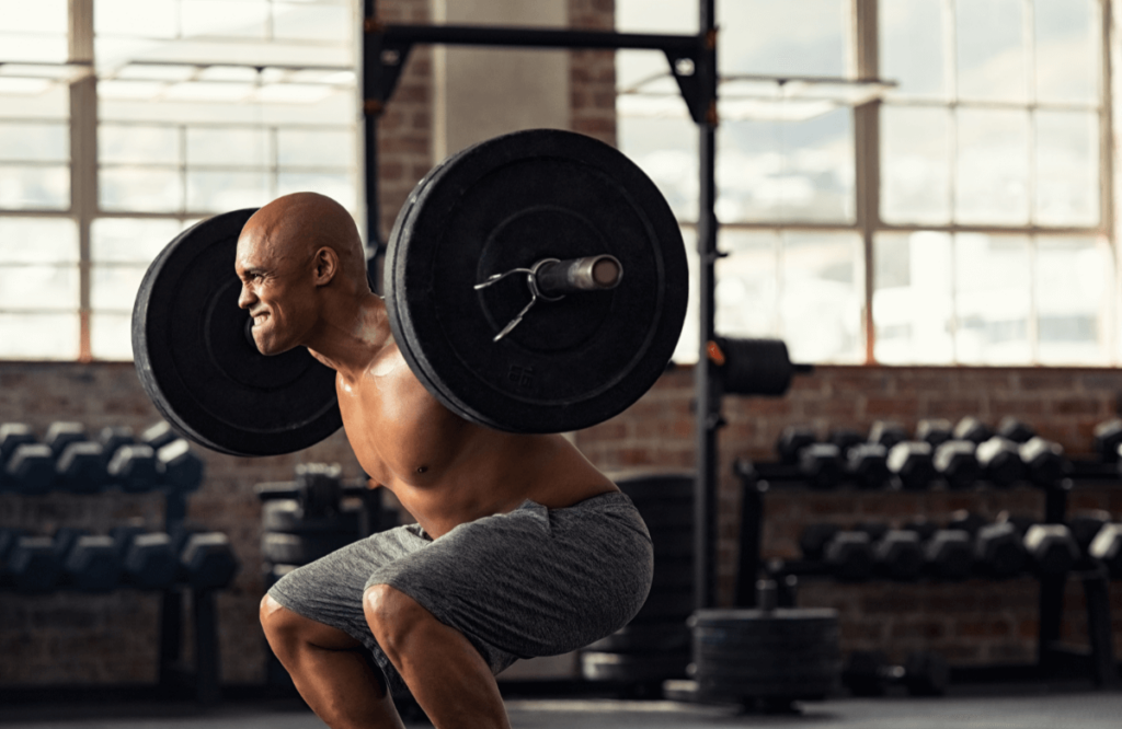 A muscular shirtless man does a back squat in a gym